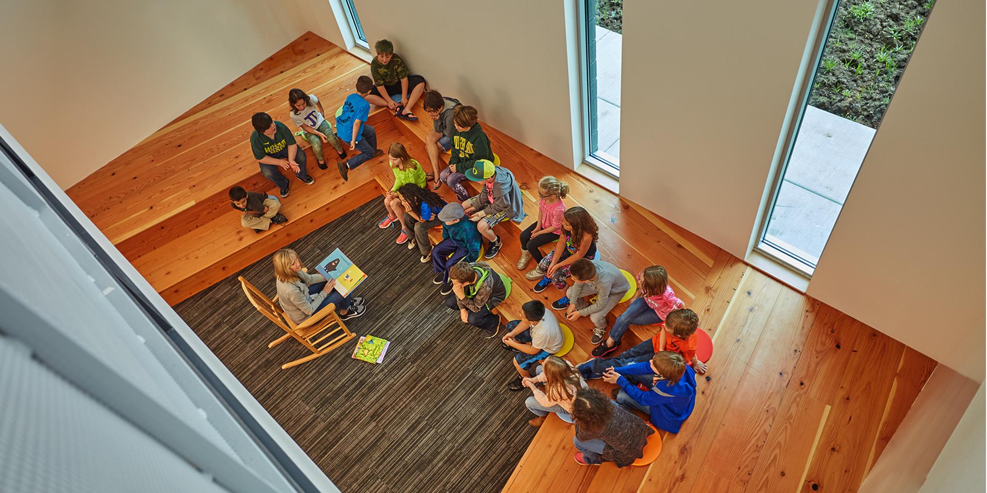 Birds eye view of students sitting and listening to teacher reading a book to the classroom